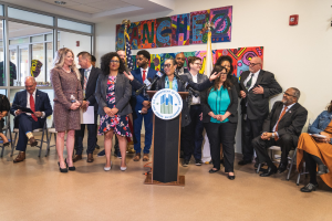 Gruppe von Menschen vor einem Podium mit Mikrofon und einer Tafel mit der Aufschrift "Sancho Stadtverordneter tritt dem Stadtverband bei", mit sitzenden und stehenden Teilnehmern, Flaggen, Feuerlöscher und Deckenbeleuchtung im Hintergrund.