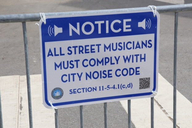 Anschlagtafel an einem Zaun mit einem Schild, auf dem "Anschlag: Alle Straßenmusiker müssen die städtische Lärmvorschrift einhalten" steht, und eine Straße im Hintergrund.