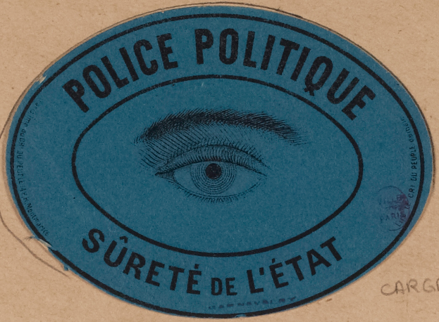 Ein brauner Karton mit einem blauen Aufkleber, der "Polizei Politique Surete de L'Etat" und ein Auge in der Mitte zeigt.