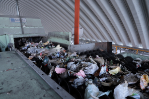 Großer Müllberg auf einem Förderband in einer Recyclinganlage mit Maschine und Metalldach im Hintergrund.