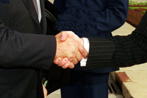 Zwei Männer im Businessanzug schütteln sich die Hände vor einem Gebäude, umgeben von grünem Gras und Pflanzen, was eine professionelle Zusammenkunft in einer förmlichen Umgebung suggeriert.