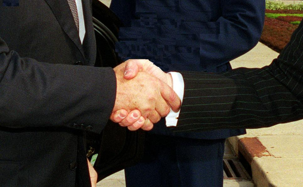 Zwei Männer im Businessanzug schütteln sich die Hände vor einem Gebäude, umgeben von grünem Gras und Pflanzen, was eine professionelle Zusammenkunft in einer förmlichen Umgebung suggeriert.