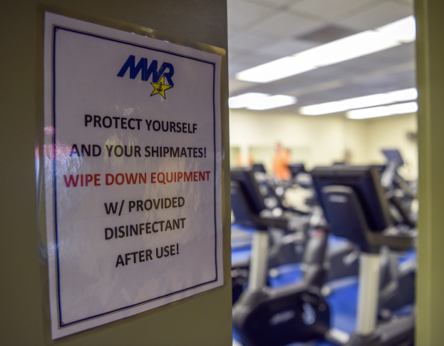 Ein Schild an einer Turnhalle mit der Aufschrift "Schützen Sie sich und Ihre Schiffskameraden" mit Laufbändern und Deckenbeleuchtung im Hintergrund, das Bild leicht unscharf, um Bewegung zu suggerieren.