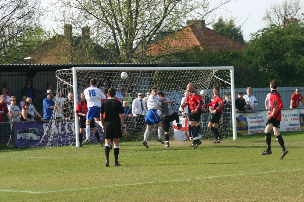 Spieler sind in ein Fußballspiel auf einem Feld mit einem Tornetz vertieft, während Zuschauer dahinter stehen, mit Bäumen und Häusern im Hintergrund.