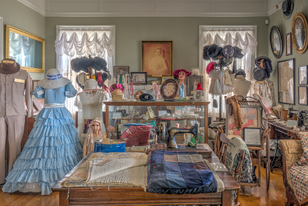 Ein Raum im Museum of Fine Arts in St. Louis, Missouri, gefüllt mit Schaufensterpuppen in verschiedenen Outfits, einem Tisch mit Kleidung und einer Nähmaschine, Stühlen mit Kissen, einem Schrank mit verschiedenen Gegenständen, Fotorahmen an der Wand, Fenstern mit Vorhängen und einer Decke.