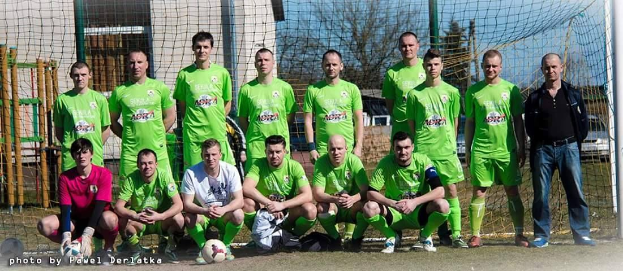 Gruppe von Männern in grünen T-Shirts auf einem Fußballfeld, einige in Hockstellung, mit einem Ball vor ihnen und einem Torpfosten rechts daneben, umgeben von Bäumen, Gebäuden und Fahrzeugen.
