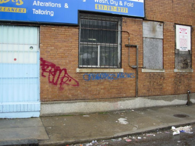 Eine Straßenansicht mit einem Gehweg und einer Straße, die mit Müll übersät sind, einer Backsteinmauer, Fenstern, Rohren, einem Tor und einer Wand am oberen Rand.