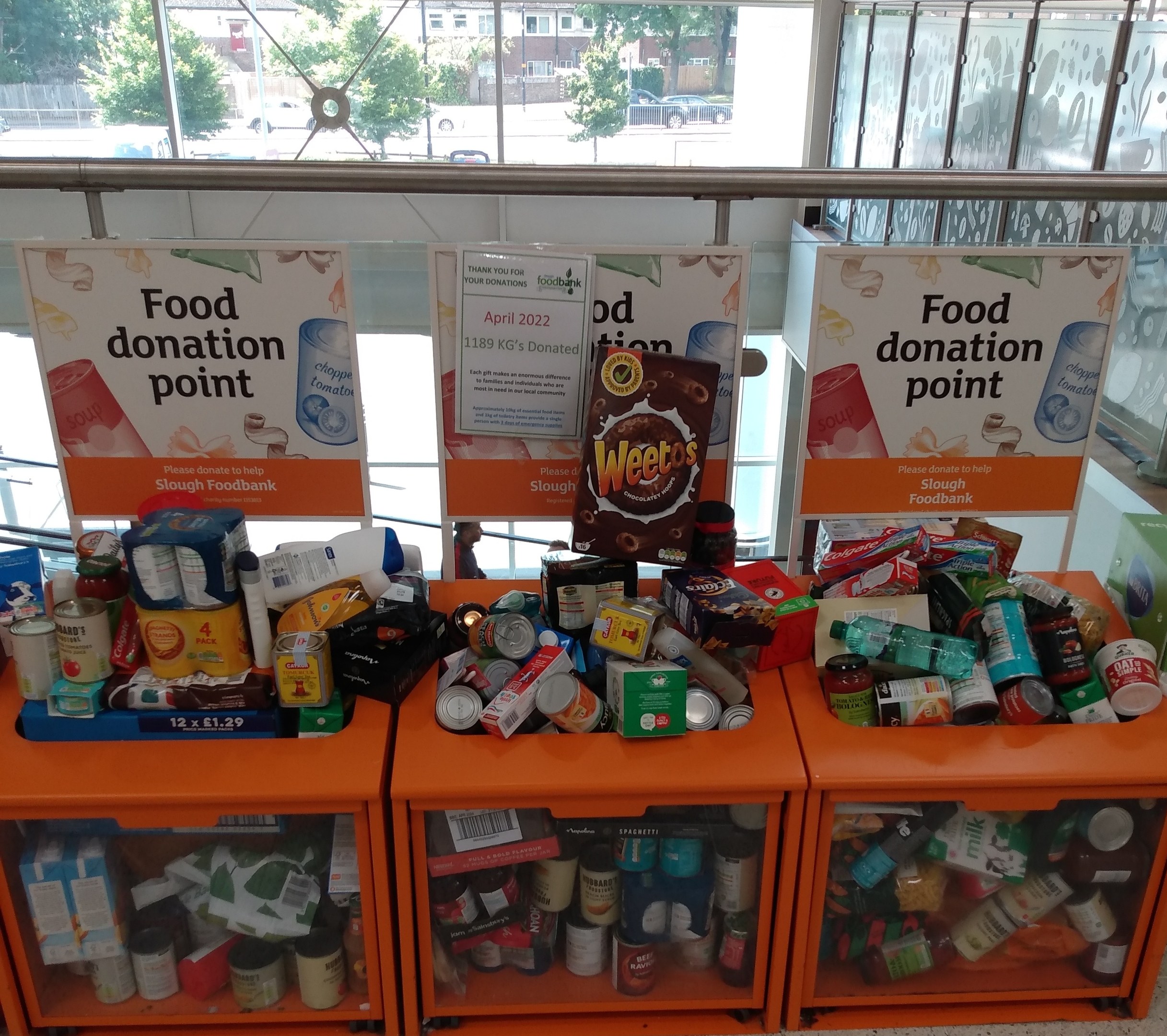 Orange Schübe mit Lebensmittelspenden gefüllt, darunter Flaschen, Dosen und Kisten, mit informativen Schildern im Hintergrund; Hintergrund zeigt Fahrzeuge, Bäume, Gebäude und Himmel durch Glasfenster.