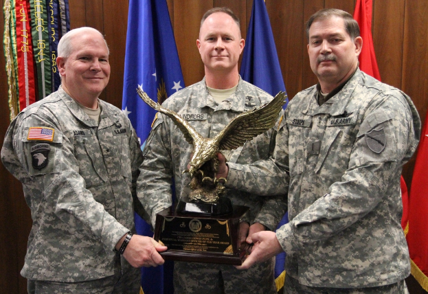 Drei Männer in Uniformen halten Auszeichnungen, mit Flaggen dahinter und buntem Text in der linken oberen Ecke.
