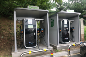 Zwei Elektroauto-Ladestationen nebeneinander, umgeben von Bäumen unter einem klaren blauen Himmel, mit einem geparkten Auto auf der rechten Seite und anderen Gegenständen auf der linken Seite in Seoul, Südkorea.