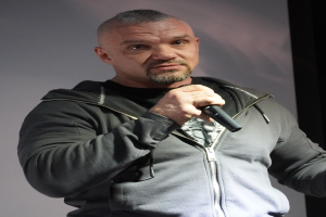 Ein Mann in einer schwarzen Kapuzenjacke und einem schwarzen Anzug mit einem weißen Hemd, zurückgekämmten Haaren und einem selbstbewussten Gesichtsausdruck hält ein Mikrofon und steht vor einer Wand.