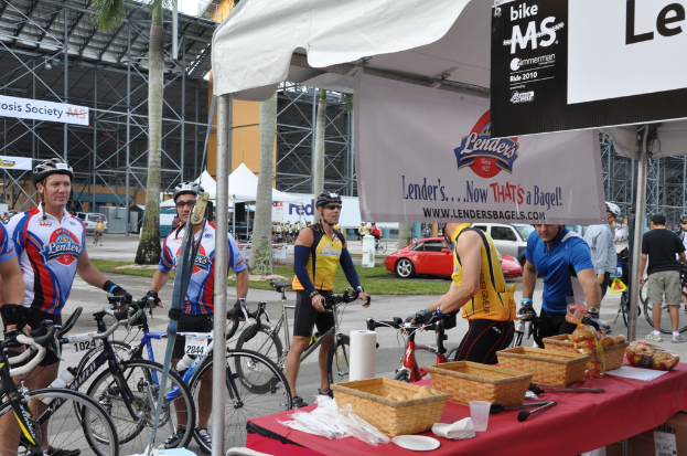 Eine Straßenszene mit Menschen in der Nähe von Fahrrädern, einem Geschäft mit Gegenständen auf einem Tisch, Bäumen, Zelten, an einen Schuppen angebrachten Bannern, Fahrzeugen und Gras.