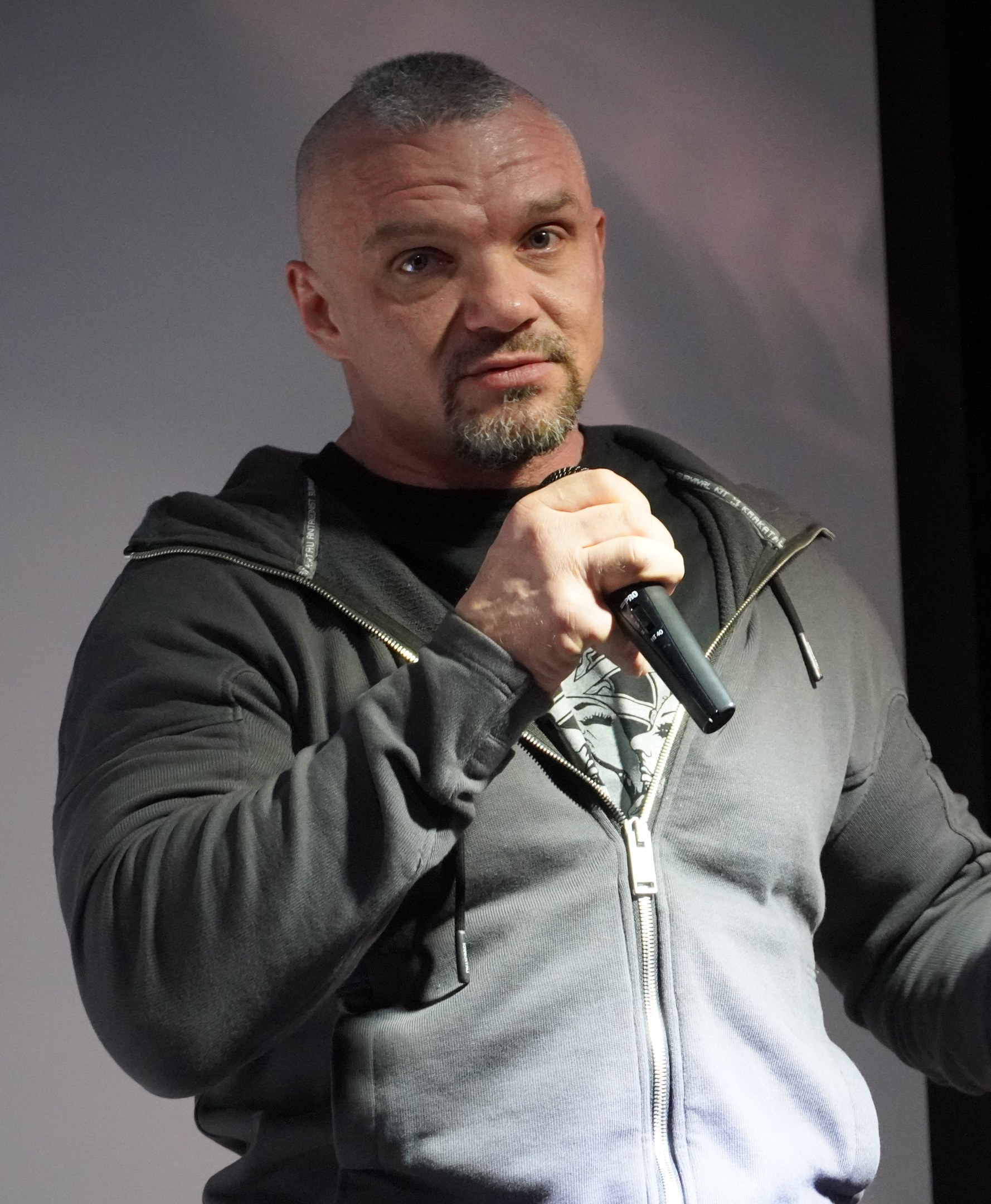 Ein Mann in einer schwarzen Kapuzenjacke und einem schwarzen Anzug mit einem weißen Hemd, glatt zurückgekämmtem Haar und einem selbstbewussten Gesichtsausdruck hält ein Mikrofon, während er vor einer Wand steht.