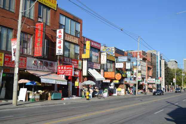 Eine belebte Stadtstraße mit hohen Gebäuden, Straßenlaternen, Strommasten, Fahrzeugen, Fußgängern und Bäumen unter einem klaren blauen Himmel.