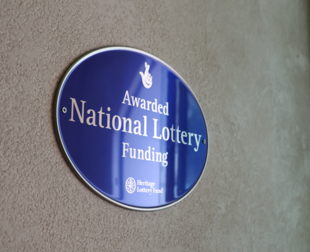 Blauer rechteckiger Schild an einem Gebäude mit weißem Hintergrund und schwarzer fettgedruckter Schrift, die "Mit Nationaler Lotterie-Förderung ausgezeichnet" lautet.