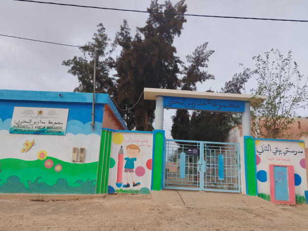 Blau-weißes Gebäude mit Wandgemälde, das als Eingang zu einer Kinder Schule dient, mit einem Tor, einem Namensschild, benachbarten Häusern, Bäumen, einem Strommast mit Drähten und einem bewölkten Himmel.