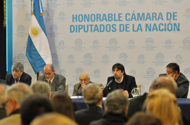 Eine Gruppe von Menschen sitzt an Tischen mit einem Krug, Papieren und einem Stift, während eine Person in der Mitte in ein Mikrofon spricht, mit einer weißen und blauen Flagge und einem Hintergrund, auf dem "ehrenwerte kamara de" steht.