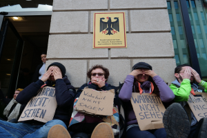 Eine Gruppe von Menschen sitzt vor einem Gebäude und hält Schilder hoch und sieht aus, als würde sie protestieren, wobei einige ihr Gesicht mit den Händen bedecken.