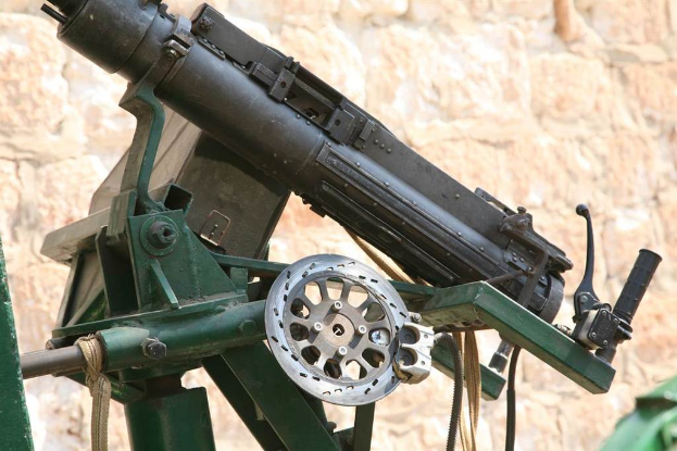 Maschinengewehr auf einem Stahlständer mit einem Griff auf der rechten Seite, Seilen und Kabeln unten und einer Wand im Hintergrund.
