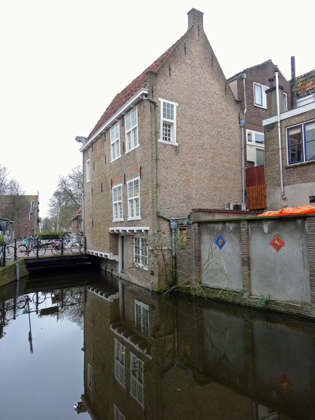 Ein Kanal in Delft, Niederlande, mit einem Gebäude auf der rechten Seite, parkenden Fahrrädern auf der linken Seite, Bäumen im Hintergrund und einem klaren blauen Himmel darüber.
