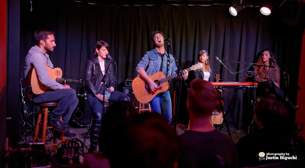 Fünf Personen auf der Bühne, zwei spielen Gitarren, alle mit Mikrofonen auf Ständern, mit einem Vorhang und Lichtern im Hintergrund.