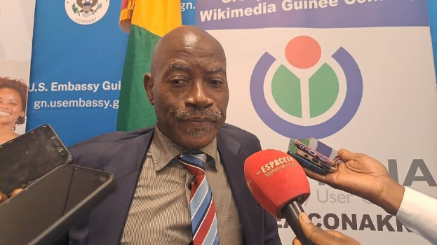 Ein Mann im Anzug spricht mit Reportern, während im Vordergrund ein Mikrofon und ein Gerät zu sehen sind, vor einer Kulisse aus Transparenten, Flaggen und einer Frau, die über Technologie in Guinea-Bissau spricht.