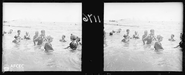 Ein Schwarz-Weiß-Foto einer Gruppe von Menschen, die in ruhigem, klarem Wasser bei einem Wettbewerb schwimmen, mit Booten im Hintergrund und Text in der linken unteren Ecke.