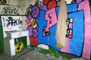 Eine beschmierte Toilette mit einem mit einem Tuch bedeckten Waschbecken und verstreuten Gegenständen auf dem Boden.