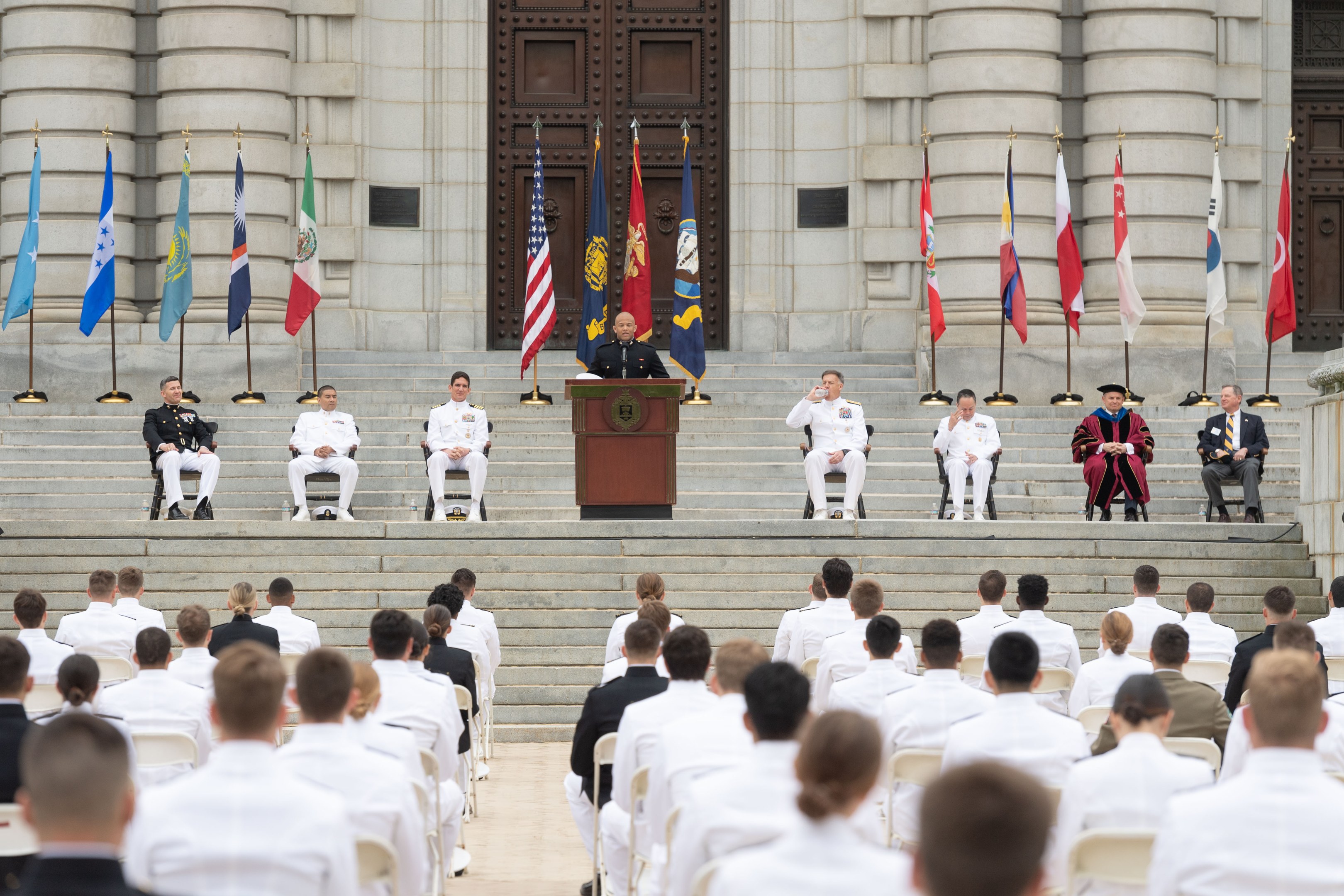 Gruppe von Menschen in weißen Uniformen auf Treppe eines Gebäudes mit einem Podium, das von Fahnen flankiert ist, vor einem großen Gebäude mit einer Tür, das eine Abschlussfeier anzeigt.