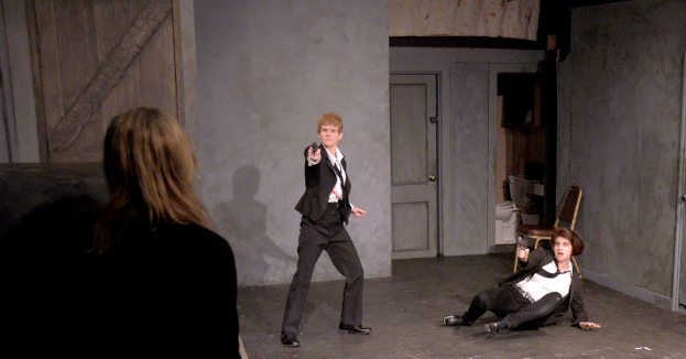 Zwei Frauen in schwarzen Anzügen, eine sitzt auf dem Boden und eine steht, beide halten Waffen, mit einer dritten Frau in der Nähe, in einem Raum mit einem Stuhl, einer Toilette und einer hölzernen Tür.