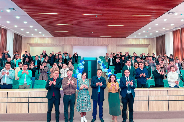 Eine Gruppe von Menschen steht vor einer Menge, einige klatschen, mit Stühlen, Tischen, Ballons, Vorhängen und Deckenleuchten bei einer Schul-Eröffnung.