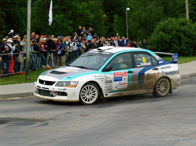 Ein Rallyefahrzeug mit Text und Logos fährt an einer Menge vorbei, die Kameras hält, mit Absperrungen, Fahnenmasten und Bäumen im Hintergrund.