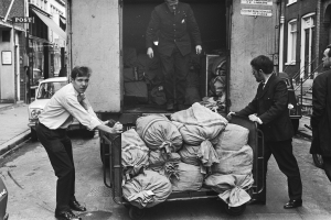 Eine Gruppe von Männern entlädt Taschen von einem Lastwagen, der auf der Seite einer Straße geparkt ist, mit einem vorbeifahrenden Auto und Gebäuden mit Fenstern, Treppen mit Geländern und Texttafeln im Hintergrund, alles in Schwarz-Weiß dargestellt.