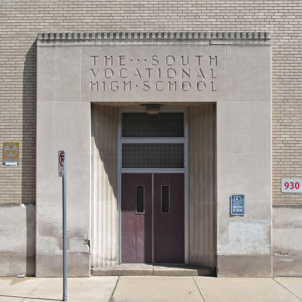 Das Südliche Berufskolleg mit einer Tür, Schilder an der Wand und einem Weg, der zur Eingangstür führt.