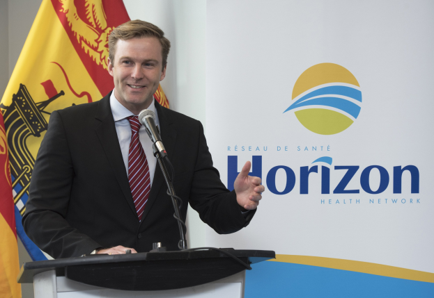 Ein Mann in einem schwarzen Blazer, weißem Hemd und roter Krawatte steht an einem Podium mit einem Mikrofon und spricht als CEO, wobei ein Banner im Hintergrund zu sehen ist.