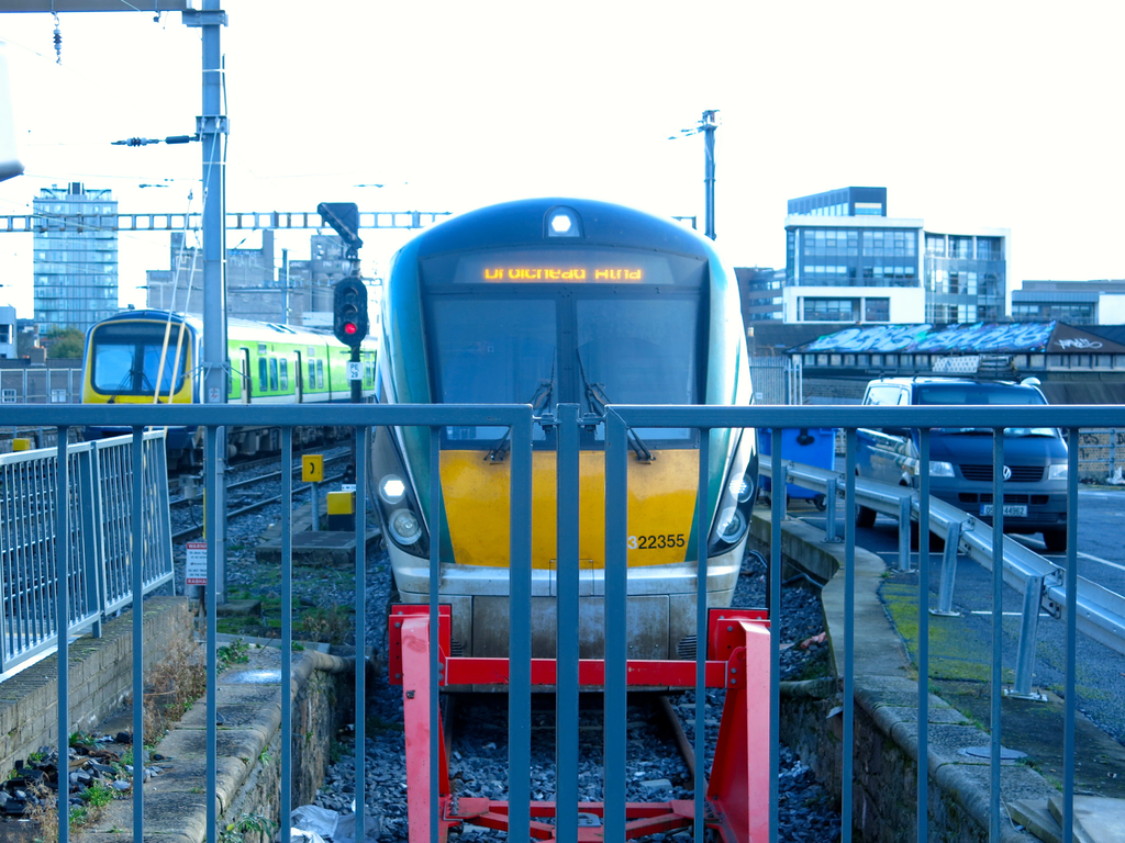 Zwei Züge auf Schienen mit einem Zaun im Vordergrund, Pfosten mit einem, der ein Signallicht hat, ein Auto auf der Straße rechts, Gebäude im Hintergrund und Himmel oben.