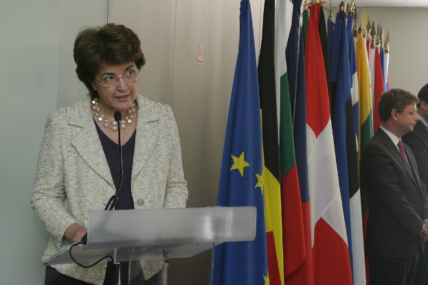 Eine Frau steht hinter einem Podium mit Mikrofon, flankiert von Flaggen, mit zwei Männern zu ihrer Rechten.