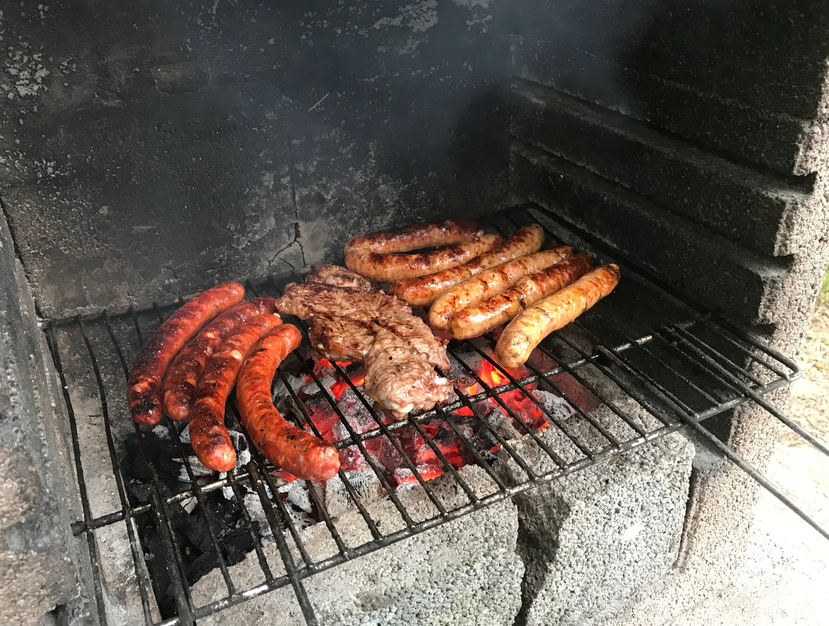 Ein Grill mit Würstchen und Fleisch, das über glühenden Kohlen gegrillt wird, mit Gras im Hintergrund.