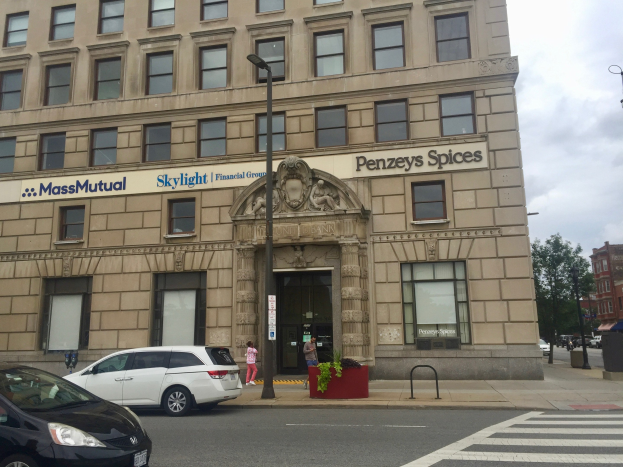 Großes Gebäude mit dem Massachusetts Mutual Financial Group-Logo, Autos vor dem Gebäude geparkt, Straßenlaternen, Lichter, fahrende Fahrzeuge, Fußgänger auf dem Gehweg, Grünflächen und ein bewölkter Himmel.