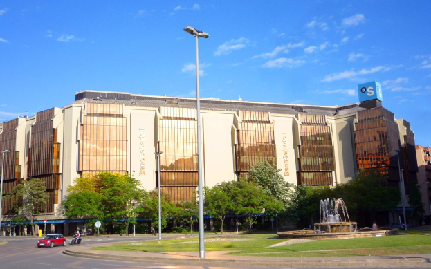 Bancroft-Stadion, ein großes Gebäude mit einem Brunnen davor, umgeben von Straßenlaternen, Autos, Fußgängern, Bäumen und einem bewölkten Himmel.