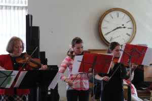 Drei Frauen auf der Bühne, die Geigen spielen, mit Notenständern und Noten davor, während ein Zuschauer im Vordergrund sitzt und zuhört; eine Uhr hängt an der weißen Wand im Hintergrund, und ein weißer Vorhang ist links zu sehen.