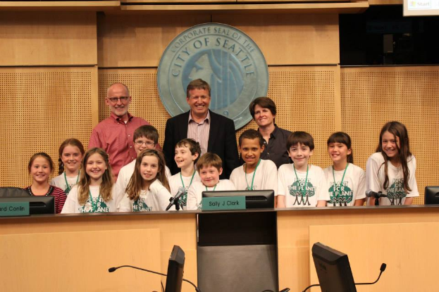 Eine Gruppe von Kindern und Erwachsenen steht vor einem Podium, lächelt und posiert für ein Foto, mit Monitoren, Mikrofonen und anderen Gegenständen auf einem Tisch im Vordergrund, einem Logo an der Wand und einem Bildschirm auf der rechten Seite im Hintergrund, bei einer Stadtratssitzung.