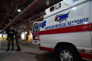 Ein Rettungswagen der American Medical Response (AMR) ist in einem Gebäude geparkt, vor ihm stehen zwei maskierte Personen; auf der linken Seite ist ein Fahrzeug zu sehen und im Hintergrund gibt es Menschen, Lichter, Rohre und andere Gegenstände.