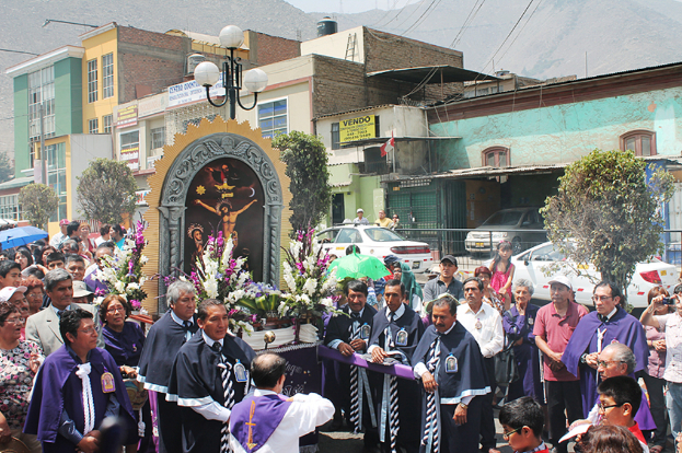Menschen auf einer Straße mit einem Kreuz und Blumengebinden in der Mitte, Autos fahren im Hintergrund und Gebäude, Bäume und Straßenlaternen im Hintergrund.