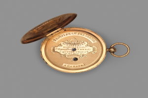 Goldene Taschenuhr mit geschlossenem Deckel auf grauem Hintergrund, mit eingraviertem Text, der wahrscheinlich die Marke oder Art angibt.