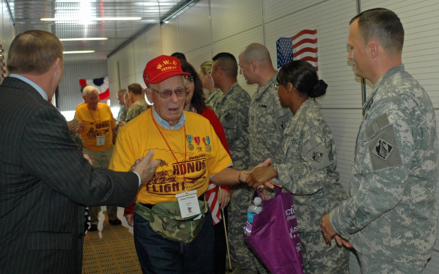 Ein belebter Flughafenvorraum mit wartenden Menschen, einige stehen und andere arbeiten, Richtungsschilder an der rechten Wand und darüber, sowie von der Decke hängende Lampen.