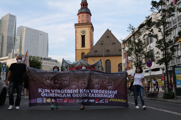 Eine Gruppe von Menschen mit Masken, die eine Straße entlanggehen und ein Banner mit Text und Bildern halten, mit einem geparkten Auto auf der linken Seite, Gebäuden, Bäumen, Schildern, Polen, einem Uhrenturm und einem klaren blauen Himmel im Hintergrund, was auf eine Protestaktion gegen Rassismus hindeutet.