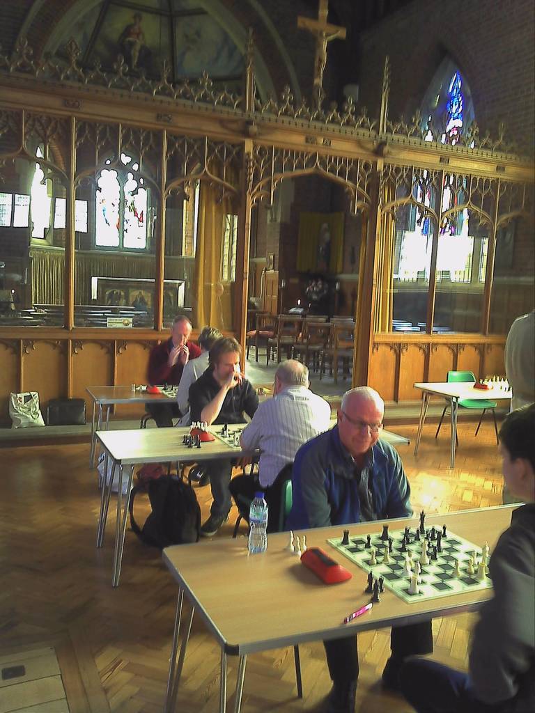 Eine Gruppe von Menschen, die an Tischen in einer Kirche Schach spielen, wobei ein Mann in einem blauen Hemd eine Flasche und einen Stift auf seinem Tisch hat und ein Glasfenster mit einem Kreuzsymbol im Hintergrund zu sehen ist.
