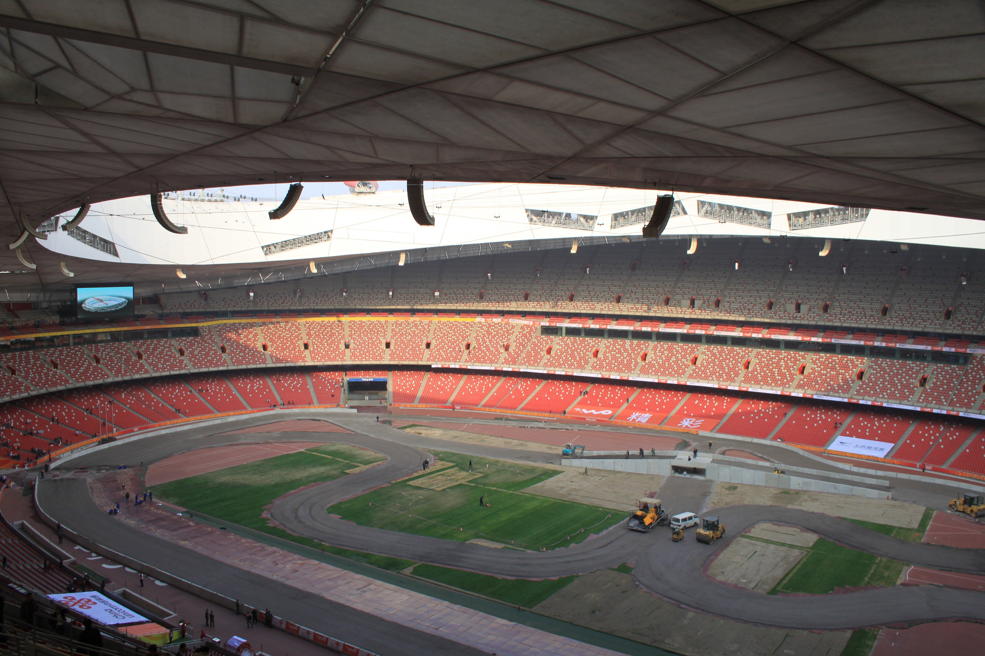 Großes Stadion voller Zuschauer, umgeben von Fahrzeugen, Gras, Texttafeln, Geländern und einem Bildschirm, mit Deckenbeleuchtung.