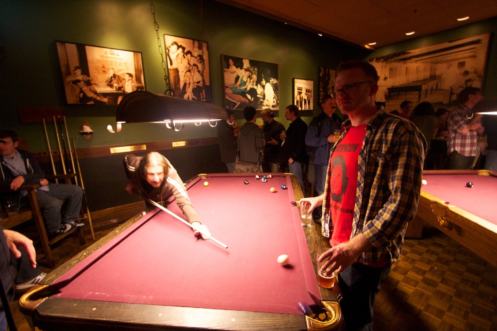 In einem Club spielen zwei Personen Snooker an einem von zwei Tischen, während mehrere andere im Hintergrund stehen und reden, vor einer cremefarbenen Wand mit Fotopostern.